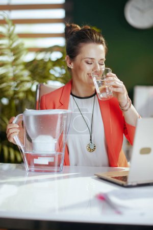 Téléchargez les photos : Un lieu de travail durable. heureuse propriétaire de petite entreprise moderne femme au travail dans une veste rouge avec tasse d'eau, filtre à eau et ordinateur portable. - en image libre de droit