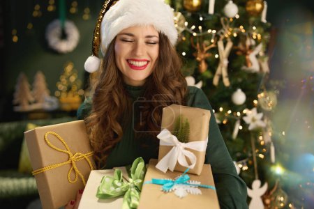 Photo for Christmas time. happy stylish woman with Santa hat and eco packaged present boxes in green dress near Christmas tree at modern home. - Royalty Free Image