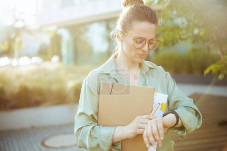 Photo for Pensive modern woman worker in business district in green blouse and eyeglasses with documents, folder and watch. - Royalty Free Image
