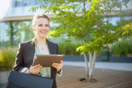 Téléchargez les photos : Heureuse femme moderne de 40 ans travailleuse près du centre d'affaires en veste noire avec tablette numérique et mallette. - en image libre de droit