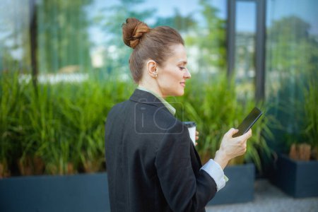 Photo for Seen from behind modern middle aged business woman near office building in black jacket with smartphone. - Royalty Free Image