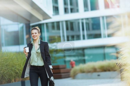 Foto de Sonriente trabajadora moderna cerca del centro de negocios en chaqueta negra con maletín y taza de café caminando. - Imagen libre de derechos