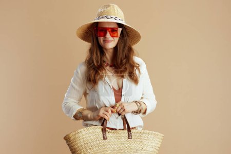 Foto de Vacaciones en playa. sonriente elegante mujer de 40 años en blusa blanca y pantalones cortos aislados en beige con bolsa de paja, sombrero de paja y gafas de sol. - Imagen libre de derechos