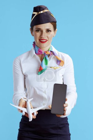 Téléchargez les photos : Hôtesse de l'air élégant femme sur fond bleu en uniforme montrant smartphone écran vide. - en image libre de droit