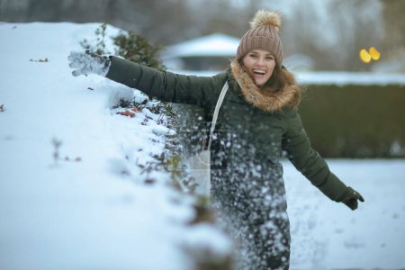 Foto de Hembra moderna feliz en abrigo verde y sombrero marrón al aire libre en el parque de la ciudad en invierno con manoplas y gorro jugando con la nieve. - Imagen libre de derechos