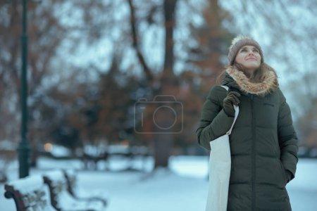 Foto de Sonriente hembra moderna en abrigo verde y sombrero marrón al aire libre en el parque de la ciudad en invierno con manoplas y gorro sombrero. - Imagen libre de derechos