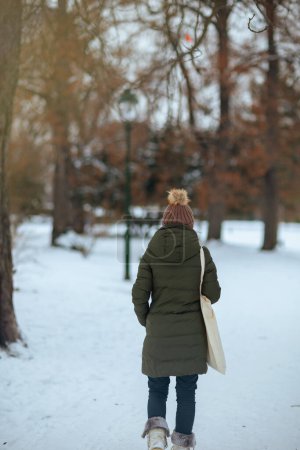 Foto de Visto desde atrás mujer moderna en abrigo verde y sombrero marrón al aire libre en el parque de la ciudad en invierno con gorro sombrero. - Imagen libre de derechos
