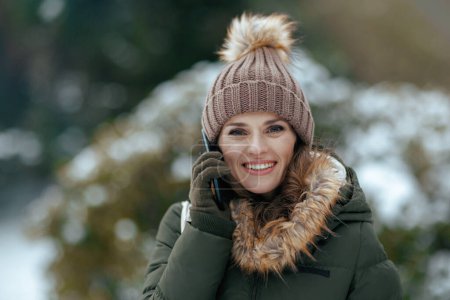 Téléchargez les photos : Femme souriante moderne de 40 ans en manteau vert et chapeau brun à l'extérieur dans le parc de la ville en hiver avec des mitaines et un bonnet parlant sur un smartphone. - en image libre de droit