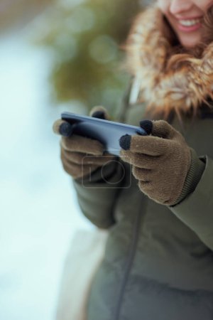 Foto de Primer plano de la mujer feliz al aire libre en el parque de la ciudad en invierno con manoplas utilizando la aplicación para teléfonos inteligentes. - Imagen libre de derechos