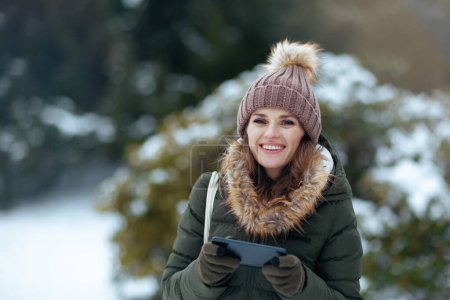 Foto de Mujer moderna feliz en abrigo verde y sombrero marrón al aire libre en el parque de la ciudad en invierno con manoplas y gorro de envío de mensajes de texto utilizando el teléfono inteligente. - Imagen libre de derechos