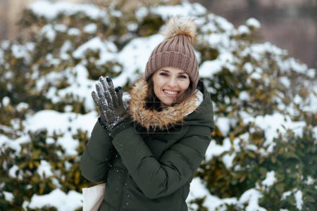 Foto de Mujer moderna feliz en abrigo verde y sombrero marrón al aire libre en el parque de la ciudad en invierno con manoplas y gorro sombrero cerca de ramas nevadas. - Imagen libre de derechos