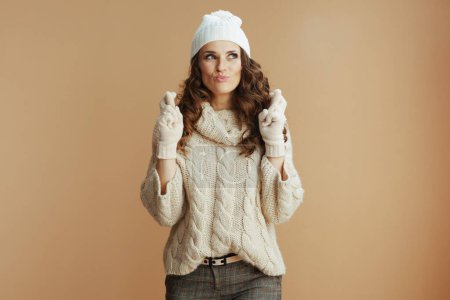 Téléchargez les photos : Bonjour l'hiver. excité femme à la mode en pull beige, mitaines et chapeau avec les doigts croisés isolé sur beige. - en image libre de droit