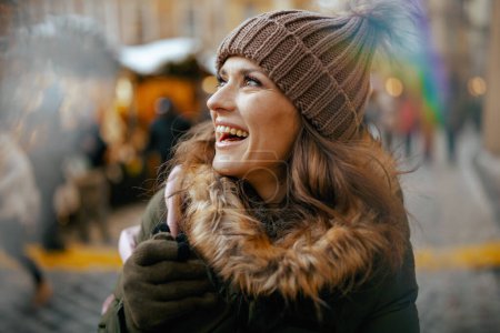 Foto de Mujer moderna feliz en abrigo verde y sombrero marrón en la feria de invierno en la ciudad mirando a la distancia. - Imagen libre de derechos