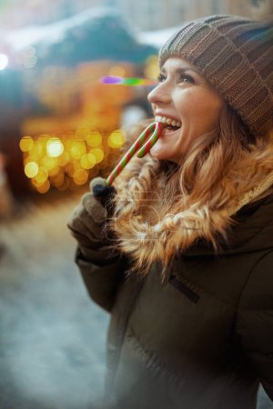 Téléchargez les photos : Heureuse jeune femme en manteau vert et chapeau brun à la foire d'hiver dans la ville avec canne à bonbons. - en image libre de droit