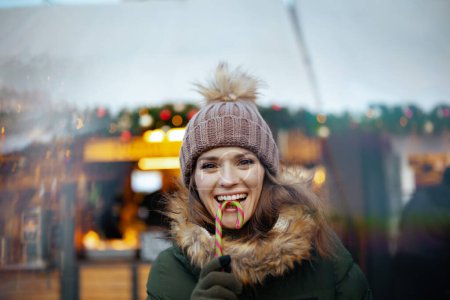 Foto de Joven sonriente con abrigo verde y sombrero marrón en la feria de invierno en la ciudad con bastón de caramelo. - Imagen libre de derechos