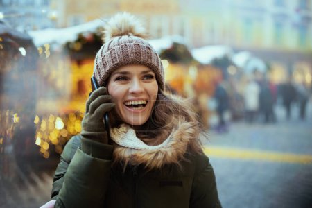 Foto de Sonriente mujer de mediana edad en abrigo verde y sombrero marrón en la feria de Navidad en la ciudad hablando en un teléfono inteligente. - Imagen libre de derechos