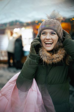 Téléchargez les photos : Femme moderne souriante en manteau vert et chapeau marron à la foire d'hiver en ville parlant sur un smartphone. - en image libre de droit