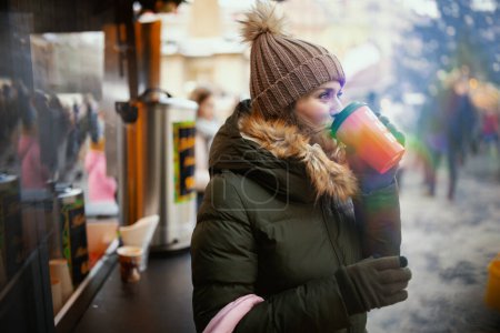 Téléchargez les photos : Moderne 40 ans femme en manteau vert et chapeau brun à la foire de Noël dans la ville avec tasse rouge de boisson chaude de Noël. - en image libre de droit