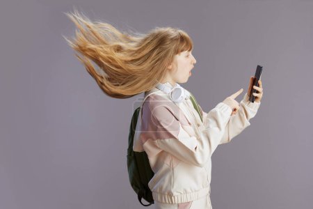 Foto de Sorprendente pupila de moda en chándal beige con mochila y auriculares usando smartphone aislado sobre fondo gris. - Imagen libre de derechos