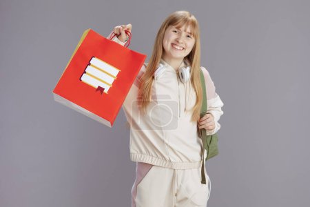 Téléchargez les photos : Portrait de heureuse écolière moderne en survêtement beige avec sac à provisions et écouteurs isolés sur gris. - en image libre de droit