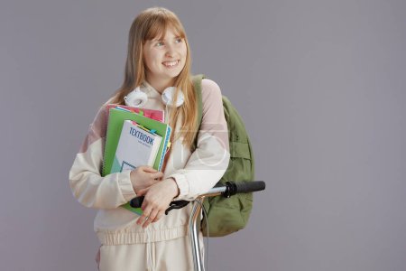 Photo for Smiling trendy teen girl in beige tracksuit with backpack, workbooks, headphones, scooter and textbook isolated on grey background. - Royalty Free Image