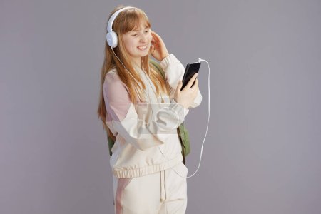 Foto de Feliz mujer joven de moda en chándal beige con teléfono inteligente escuchando la música con auriculares contra gris. - Imagen libre de derechos
