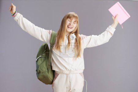 Foto de Sonriente chica adolescente moderna en chándal beige con mochila, libros de trabajo y auriculares contra gris con los brazos levantados regocijo. - Imagen libre de derechos