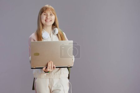 Photo for Portrait of smiling trendy young woman in beige tracksuit with headphones using laptop against grey. - Royalty Free Image