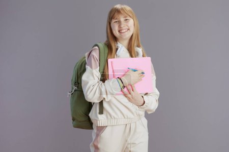 Photo for Portrait of smiling modern teenage girl in beige tracksuit with backpack and workbooks against grey. - Royalty Free Image