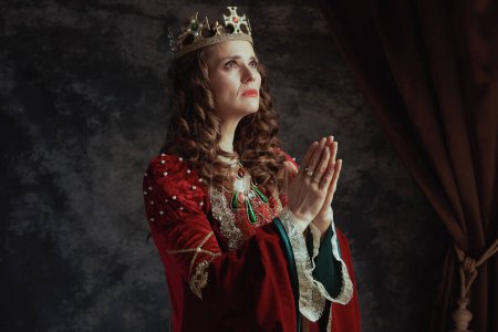 Photo for Medieval queen in red dress with crown praying on dark gray background. - Royalty Free Image