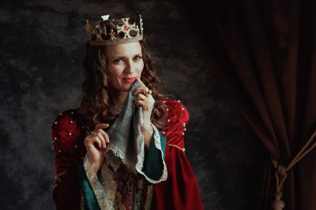 Photo for Smiling medieval queen in red dress with handkerchief and crown on dark gray background. - Royalty Free Image
