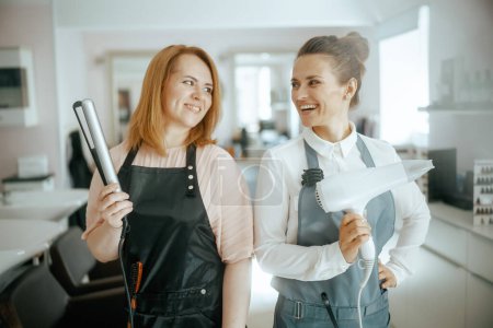 Photo for Happy two women hairdressers in uniform in modern beauty studio with hair straightener and hair dryer. - Royalty Free Image