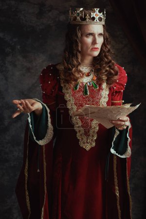 Téléchargez les photos : Reine médiévale en robe rouge avec parchemin et couronne sur fond gris foncé. - en image libre de droit