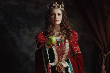 Photo for Medieval queen in red dress with green apple and crown on dark gray background. - Royalty Free Image