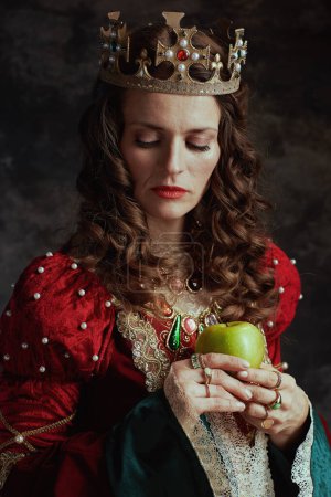 Photo for Medieval queen in red dress with green apple and crown on dark gray background. - Royalty Free Image