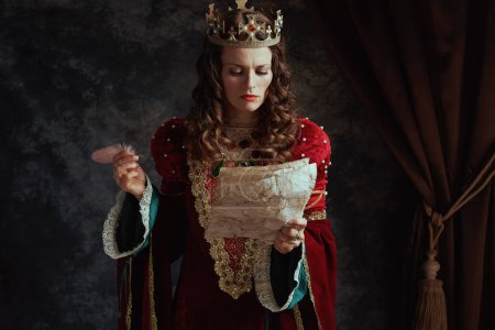 Photo for Medieval queen in red dress with parchment and crown on dark gray background. - Royalty Free Image