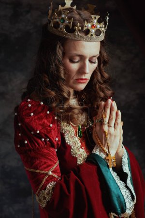 Foto de Reina medieval en vestido rojo con rosario y corona rezando sobre fondo gris oscuro. - Imagen libre de derechos