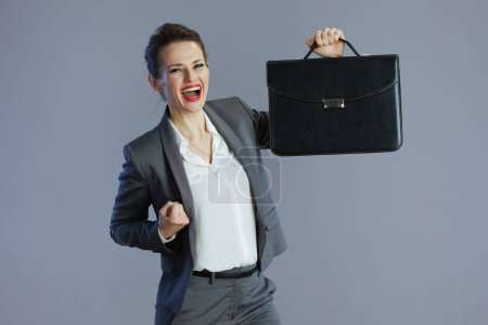 Photo for Smiling trendy 40 years old woman employee in grey suit with briefcase isolated on gray. - Royalty Free Image