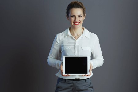 Photo for Happy trendy 40 years old woman worker in white blouse showing tablet PC blank screen isolated on grey background. - Royalty Free Image