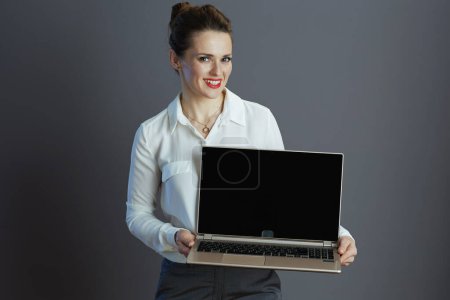 Foto de Sonriente elegante dueño de una pequeña empresa mujer en blusa blanca que muestra la pantalla en blanco portátil aislado en fondo gris. - Imagen libre de derechos