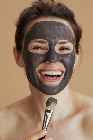 Foto de Sonriente mujer de mediana edad con cepillo y máscara cosmética sobre fondo beige. - Imagen libre de derechos