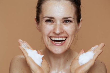 Foto de Sonriendo 40 años de edad, mujer con espuma facial limpiador lavado de la cara aislado en beige. - Imagen libre de derechos