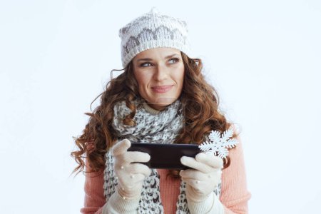 Photo for Hello winter. pensive modern 40 years old woman in sweater, mittens, hat and scarf against white background with snowflake sending text message using smartphone. - Royalty Free Image