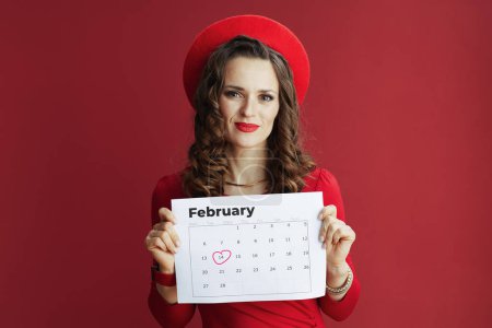 Foto de Feliz San Valentín. elegante mujer de 40 años en vestido rojo y boina sobre fondo rojo con calendario de febrero. - Imagen libre de derechos