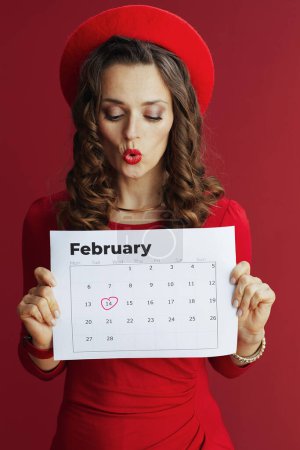 Téléchargez les photos : Joyeux Valentin. surpris femme d'âge moyen moderne en robe rouge et béret sur fond rouge avec calendrier Février. - en image libre de droit
