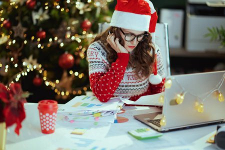 Photo for Christmas time. tired stylish small business owner woman in santa hat in eyeglasses in red Christmas sweater with documents and laptop working in modern green office with Christmas tree. - Royalty Free Image