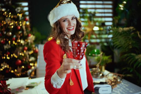 Téléchargez les photos : Noël. souriant élégante propriétaire de petite entreprise d'âge moyen femme en santa chapeau et veste rouge avec verre à vin dans le bureau vert moderne avec arbre de Noël. - en image libre de droit