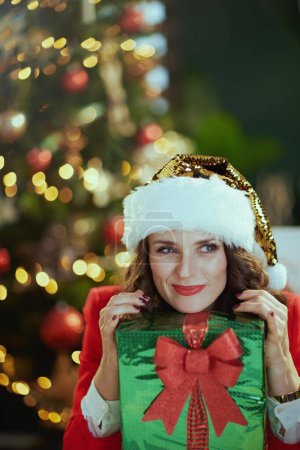 Foto de En Navidad. pensativo elegante en chaqueta roja con caja actual en sombrero de santa en moderna oficina verde con árbol de Navidad. - Imagen libre de derechos