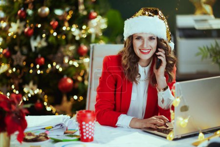 Téléchargez les photos : Noël. souriant élégante propriétaire de petite entreprise d'âge moyen femme en santa chapeau et veste rouge avec ordinateur portable parlant sur un smartphone et travaillant dans un bureau vert moderne avec arbre de Noël. - en image libre de droit