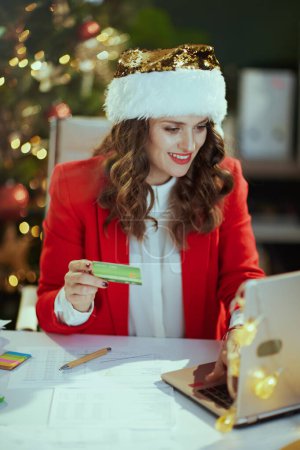 Photo for Christmas time. smiling elegant middle aged business woman in santa hat and red jacket with laptop and credit card in modern green office with Christmas tree. - Royalty Free Image